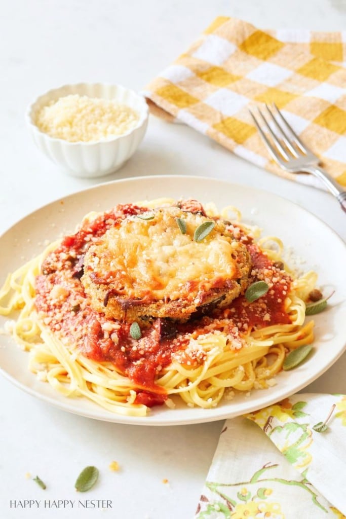 A plate of spaghetti topped with tomato sauce and breaded, cheese-covered eggplant slices makes for delicious dinner ideas. Grated cheese is sprinkled on top, with a yellow checkered napkin, two forks, and a small bowl of grated cheese nearby.