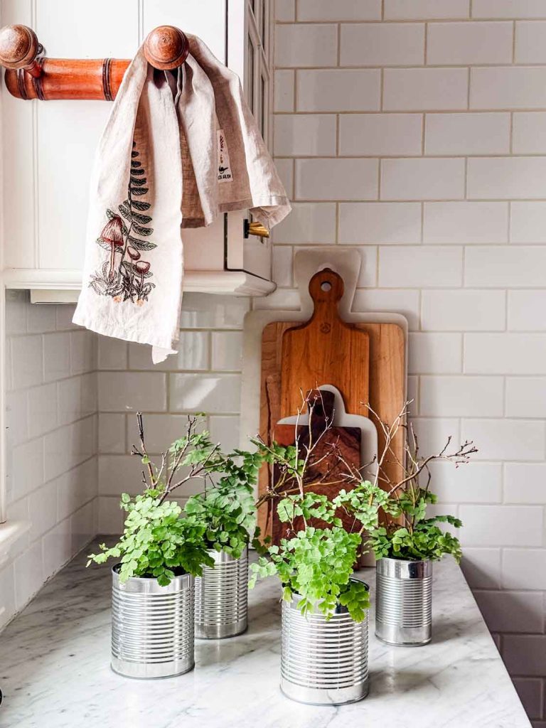 Three tin cans used as plant pots with green leafy plants sit on a marble countertop. Behind them, wooden cutting boards are stacked upright, and a tea towel hangs from a wooden rack on white tiled kitchen walls.