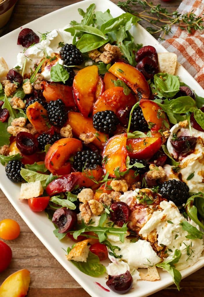 A rectangular platter of salad with fresh arugula, grilled peach slices, blackberries, cherries, crumbled cheese, and a crunchy topping, garnished with herbs—a vibrant option for your summer dinner ideas.