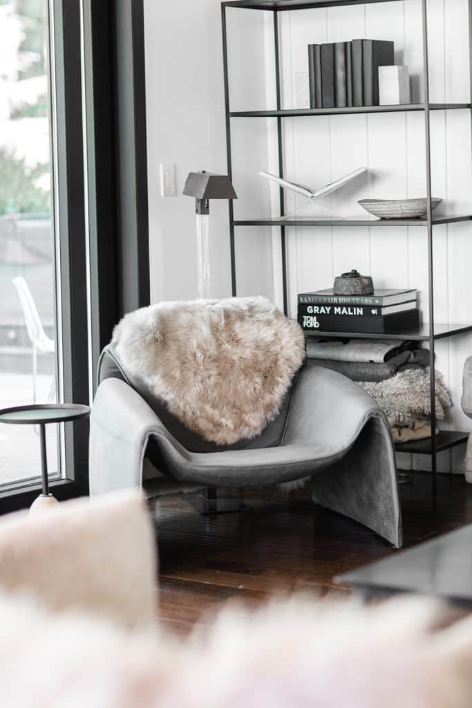 A modern gray lounge chair with a beige faux fur throw sits by a large window. Behind it, a black metal bookshelf holds books and decor. Sunlight brightens the cozy, minimalist room with wood floors.