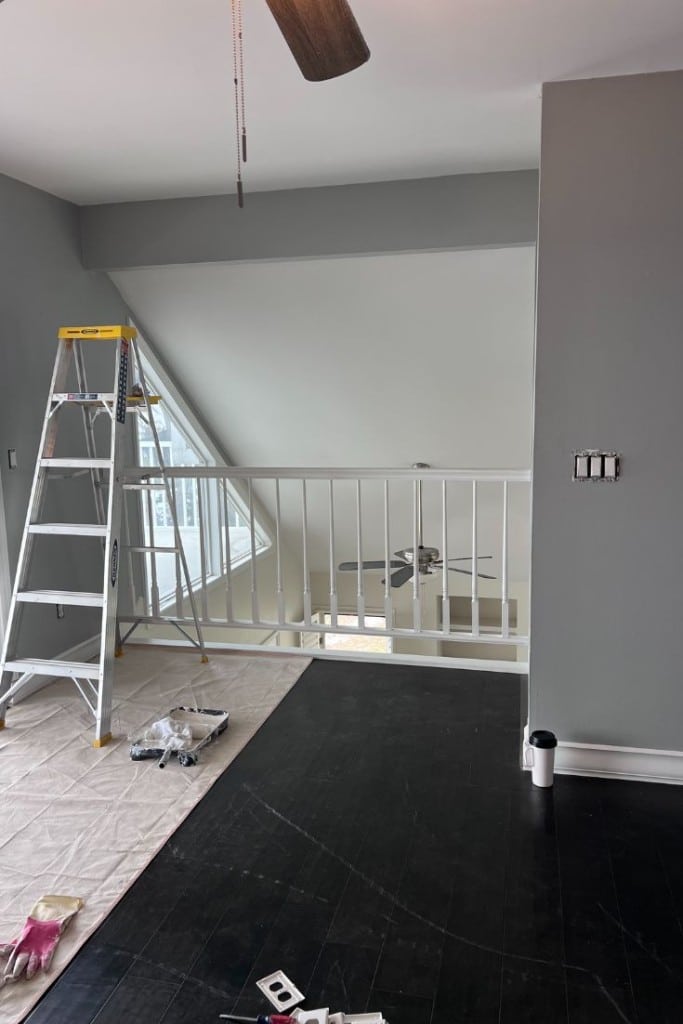A room with dark wood floors, a gray wall, and a white railing overlooking a loft with large windows. A ladder, drop cloth, and painting supplies are set up, indicating ongoing renovations. A ceiling fan is visible below.