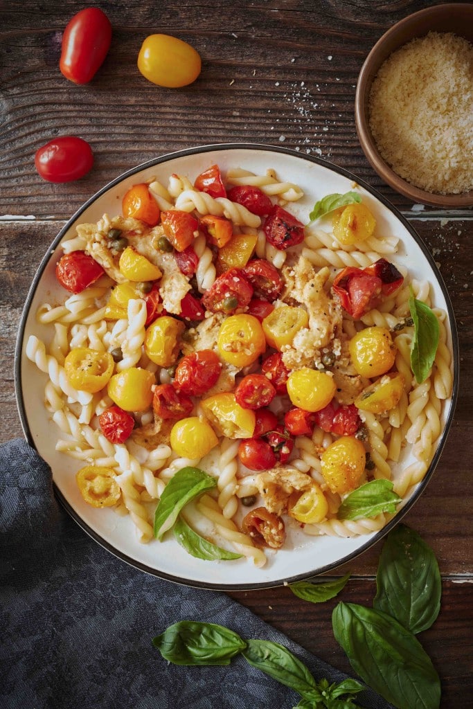 A plate of pasta topped with roasted red and yellow cherry tomatoes, chicken pieces, fresh basil leaves, and grated cheese—one of the tastiest dinner ideas—set on a rustic wooden table with a bowl of cheese and basil nearby.