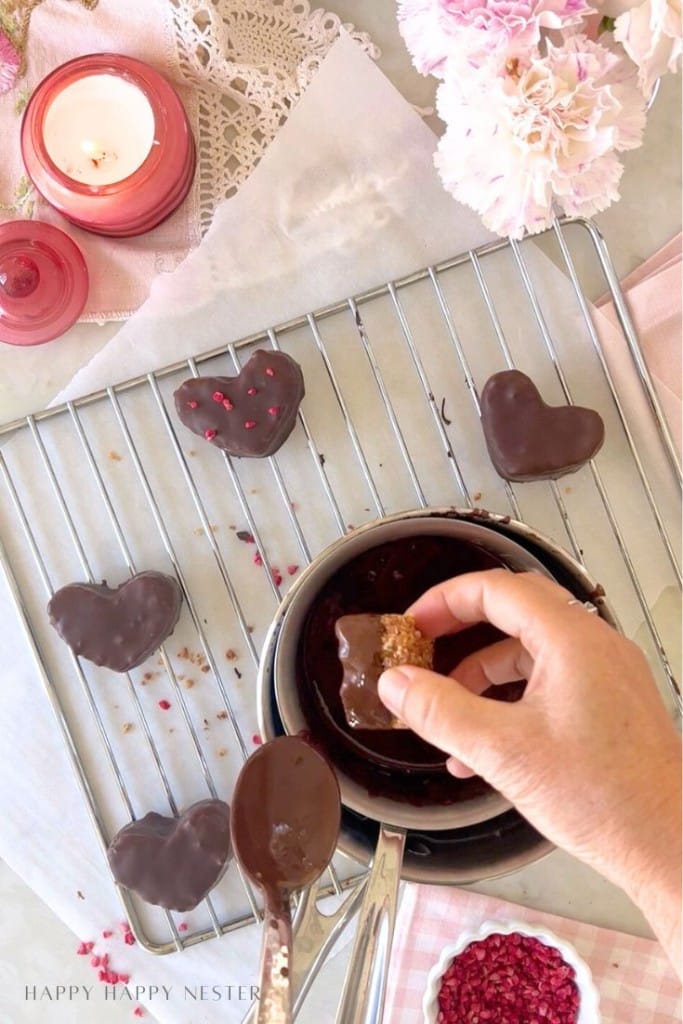 A hand dips a heart-shaped treat into melted chocolate over a pot, with more chocolate covered quinoa recipe treats cooling on a rack. Pink flowers, a candle, and pink decorations add charm to the table.