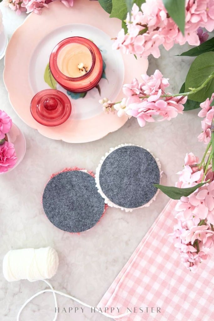 A flat lay of two round felt coasters, a candle, pink flowers, a spool of white string, and a pink gingham cloth on a marble surface. The scene is bright and decorative.
