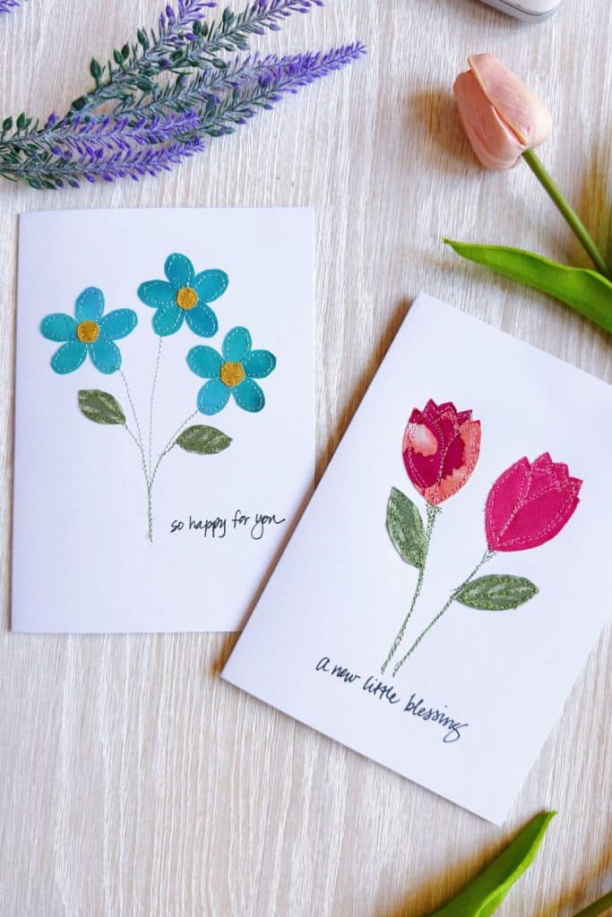 Two greeting cards on a light wooden surface: one with punch needle blue flowers and “so happy for you,” the other with two red tulips and “a new little blessing.” DIY purple and pink blooms and green leaves surround them.