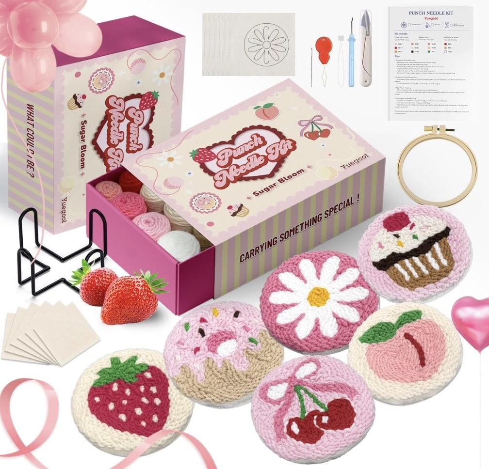 A punch needle crafting kit with tools, yarn, fabric, a wooden hoop, instructions, and six finished embroidered designs: daisy, cupcake, macaron, donut, strawberry, and cherries—all pastel on a pink background for DIY fun.