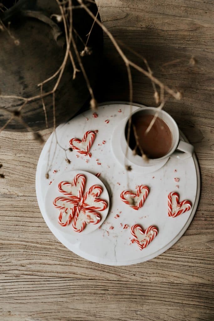 A white mug of hot chocolate sits on a round marble board, surrounded by red and white candy canes in heart and flower shapes&mdash;perfect inspiration for your next chocolate covered quinoa recipe&mdash;on a rustic wooden surface with dried plant stems above.