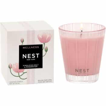 A pink scented candle labeled "NEST New York" sits beside its box, which is decorated with pink lotus flowers and labeled "Wellness Himalayan Salt & Rosewater." The candle flame is lit.