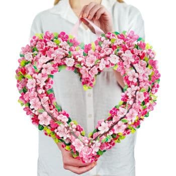 A person in a white shirt holds a large, heart-shaped wreath made of pink and white artificial flowers and green leaves. The heart wreath is vibrant and colorful, with a ribbon for hanging.