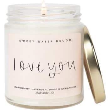 A lit candle in a glass jar with a gold lid resting beside it. The label reads "I love you" and lists the scents: mahogany, lavender, wood, and geranium. Brand: Sweet Water Decor.