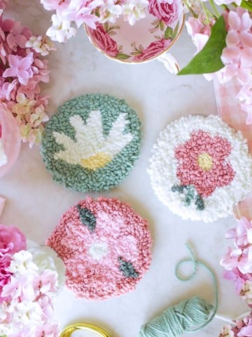 Three round, handmade punch needle coasters with floral designs are arranged on a white surface, surrounded by pink flowers, a floral teacup, green yarn, and punch needle crafting DIY supplies.