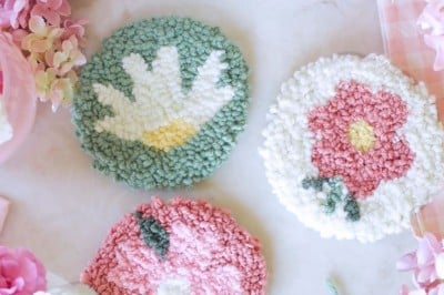 Three round, handmade punch needle coasters with floral designs are arranged on a white surface, surrounded by pink flowers, a floral teacup, green yarn, and punch needle crafting DIY supplies.