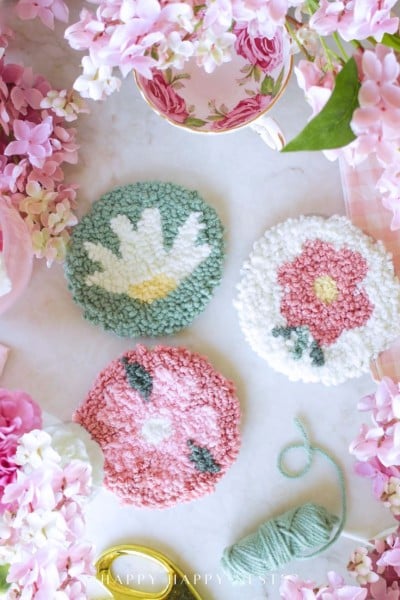 Three round, handmade punch needle coasters with floral designs are arranged on a white surface, surrounded by pink flowers, a floral teacup, green yarn, and punch needle crafting DIY supplies.
