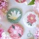 Three round, handmade punch needle coasters with floral designs are arranged on a white surface, surrounded by pink flowers, a floral teacup, green yarn, and punch needle crafting DIY supplies.
