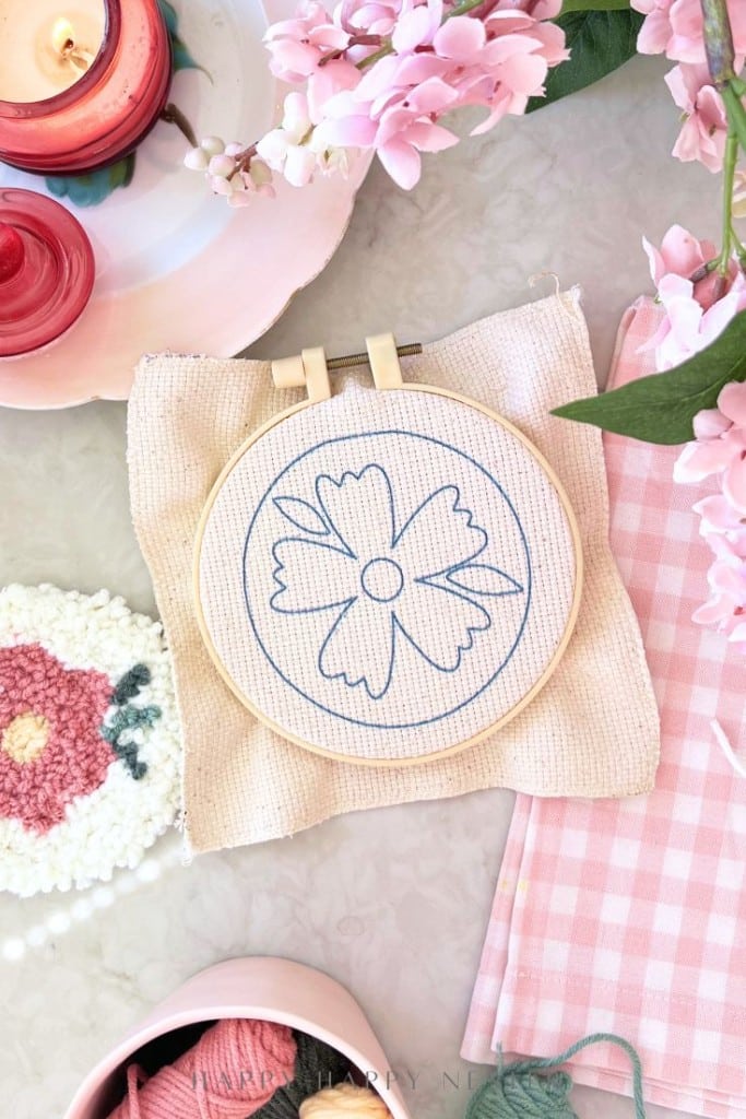 An embroidery hoop with fabric displays a simple blue outline of a flower. Surrounding it are pink flowers, a pink gingham cloth, candles, and a small round floral coaster on a white surface.