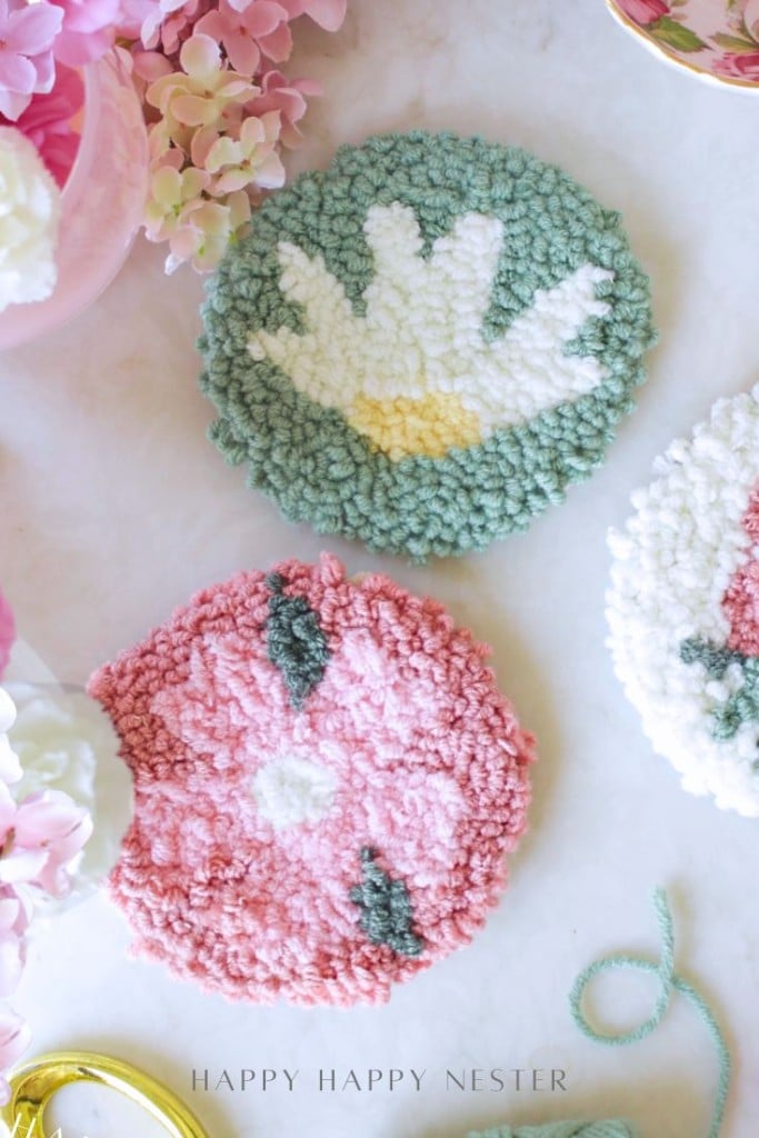 Two round, handmade tufted coasters with floral designs—one pink with a white and green flower, the other green with a white and yellow flower—lie on a white surface surrounded by pink flowers.