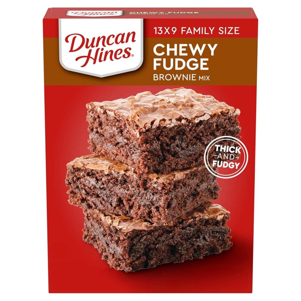 Box of Duncan Hines Chewy Fudge Brownie Mix, 13x9 family size, with an image of three stacked, thick, fudgy brownies on the front. Perfect for your next heart shaped brownies recipe. Red background with "Thick and Fudgy" in a white circle.