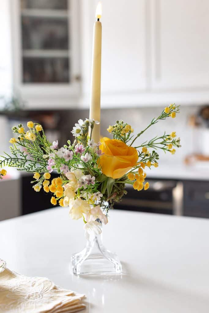 A lit cream-colored taper candle in a glass holder is decorated with yellow roses, small white and yellow flowers, and greenery—a charming DIY crafting accent for your white kitchen countertop.