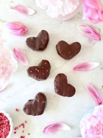 Six heart-shaped chocolate-covered cookies arranged on a white surface, surrounded by pink flower petals and blooms, with pink sprinkles nearby, inspire the perfect treat or even a Chocolate Covered Quinoa Recipe for a fun twist.