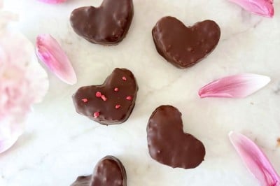 Six heart-shaped chocolate-covered cookies arranged on a white surface, surrounded by pink flower petals and blooms, with pink sprinkles nearby, inspire the perfect treat or even a Chocolate Covered Quinoa Recipe for a fun twist.