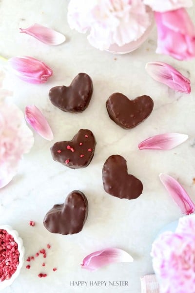 Six heart-shaped chocolate-covered cookies arranged on a white surface, surrounded by pink flower petals and blooms, with pink sprinkles nearby, inspire the perfect treat or even a Chocolate Covered Quinoa Recipe for a fun twist.