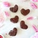 Six heart-shaped chocolate-covered cookies arranged on a white surface, surrounded by pink flower petals and blooms, with pink sprinkles nearby, inspire the perfect treat or even a Chocolate Covered Quinoa Recipe for a fun twist.