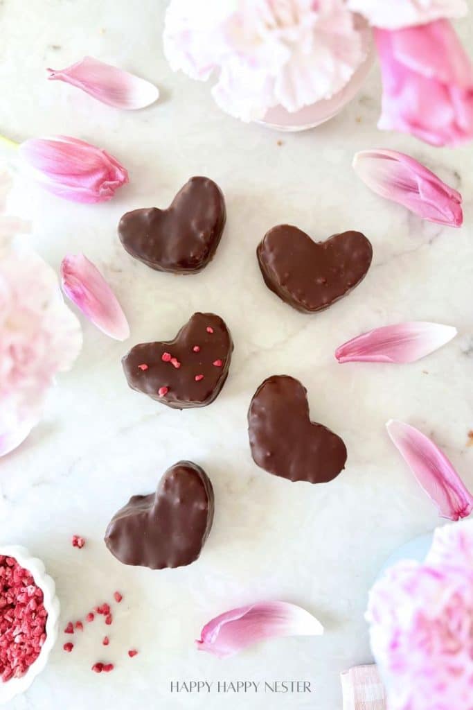 Six heart-shaped chocolate-covered treats, inspired by a chocolate covered quinoa recipe, are arranged on a white surface with pink flower petals and bits of red garnish. The scene is bright and romantic, with soft flowers in the background.