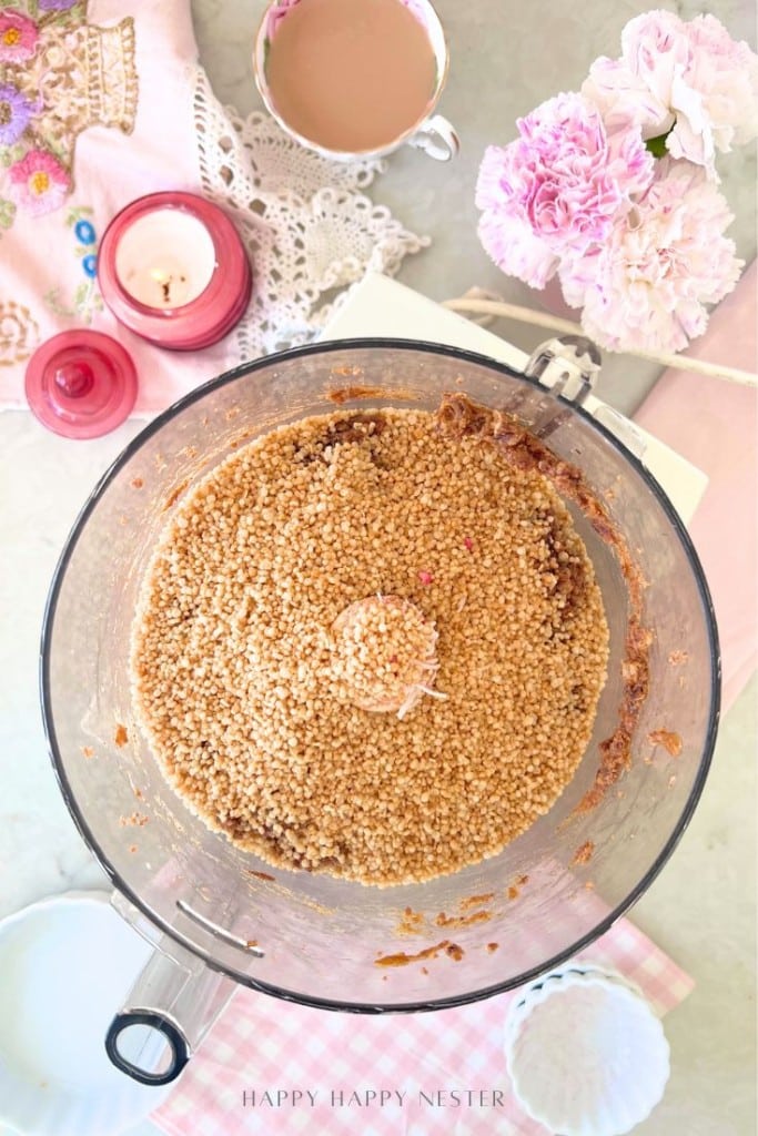 A food processor filled with a crumbly mixture sits on a light surface surrounded by a pink candle, a cup of coffee, pink flowers, a white dish, and a notebook with a pink cover.