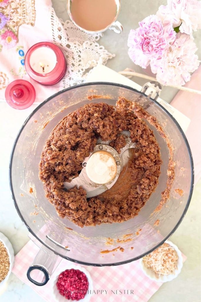 A food processor contains a thick brown mixture. Surrounding it are pink candles, a teacup, flowers, and small bowls of seeds and dried fruit, all arranged on a pastel tablecloth for a cozy baking setup.