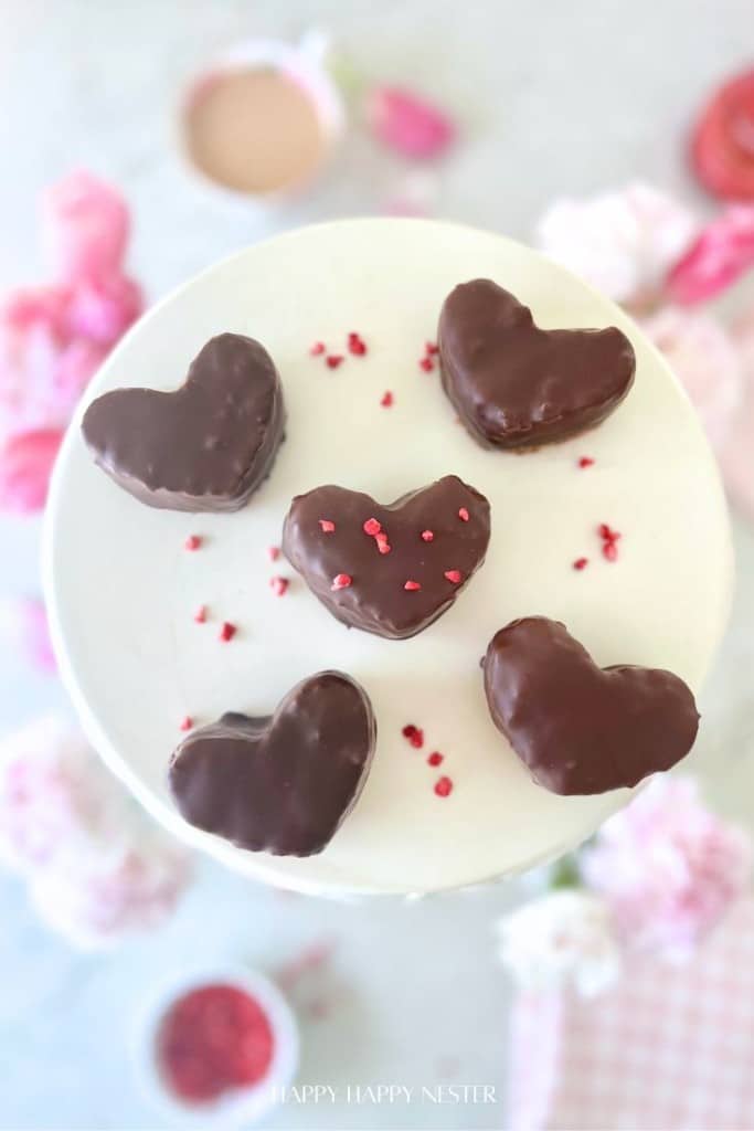 Five heart-shaped chocolate-covered treats, inspired by a Chocolate Covered Quinoa Recipe, are arranged on a white cake stand. One heart is decorated with small pink sprinkles, set against soft pink and white flowers and blurred baking items.