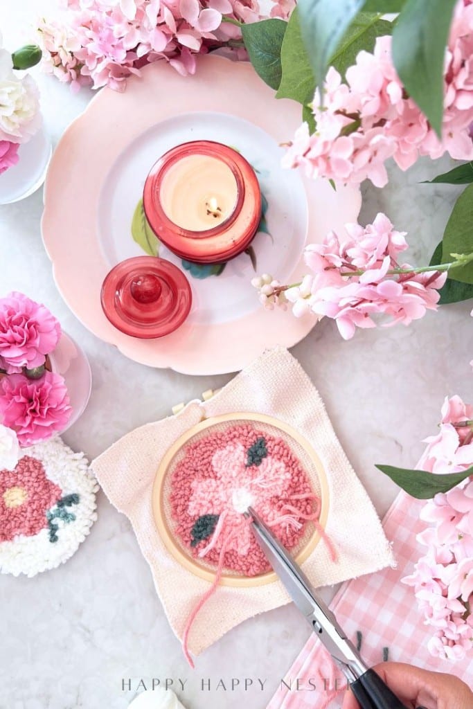 A pink-themed flat lay features a candle, pink flowers, and an embroidery hoop with a floral pattern, surrounded by scissors and other sewing supplies on a white surface.