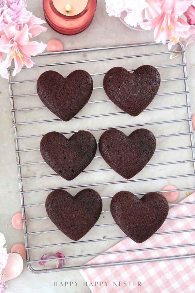 Six heart-shaped chocolate cakes are cooling on a wire rack, surrounded by pink flowers, a pink candle, and a pink gingham cloth, creating a romantic and festive atmosphere.