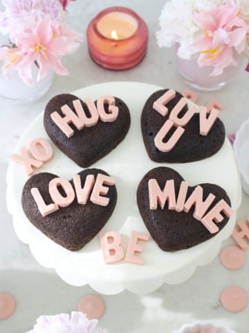A white cake stand holds four heart-shaped chocolate cakes made with a heart shaped brownies recipe, each topped with pink letters spelling &ldquo;HUG,&rdquo; &ldquo;LUV U,&rdquo; &ldquo;LOVE,&rdquo; and &ldquo;MINE.&rdquo; Pink candies, flowers, and a lit candle complete the romantic setting.