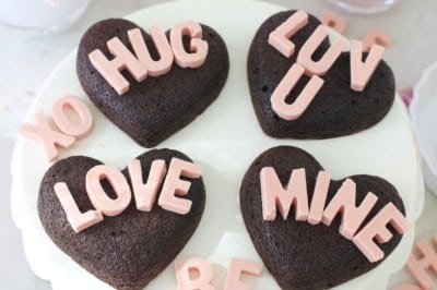 A white cake stand holds four heart-shaped chocolate cakes made with a heart shaped brownies recipe, each topped with pink letters spelling “HUG,” “LUV U,” “LOVE,” and “MINE.” Pink candies, flowers, and a lit candle complete the romantic setting.