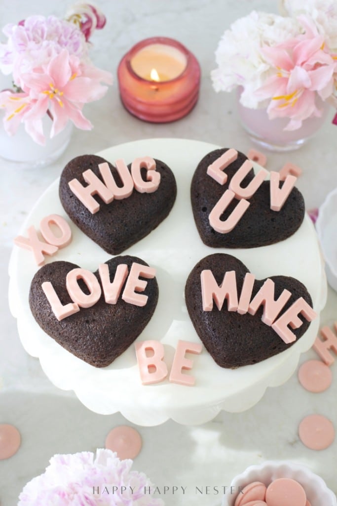 A white cake stand holds four heart-shaped chocolate cakes made with a heart shaped brownies recipe, each topped with pink letters spelling “HUG,” “LUV U,” “LOVE,” and “MINE.” Pink candies, flowers, and a lit candle complete the romantic setting.