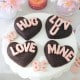 A white cake stand holds four heart-shaped chocolate cakes made with a heart shaped brownies recipe, each topped with pink letters spelling &ldquo;HUG,&rdquo; &ldquo;LUV U,&rdquo; &ldquo;LOVE,&rdquo; and &ldquo;MINE.&rdquo; Pink candies, flowers, and a lit candle complete the romantic setting.