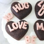 Heart-shaped chocolate cakes, inspired by a heart shaped brownies recipe, spell out phrases like "LOVE," "HUG," and "XO" in pink letters, all arranged on a white cake stand with a pink flower nearby.