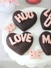 Heart-shaped chocolate cakes, inspired by a heart shaped brownies recipe, spell out phrases like "LOVE," "HUG," and "XO" in pink letters, all arranged on a white cake stand with a pink flower nearby.