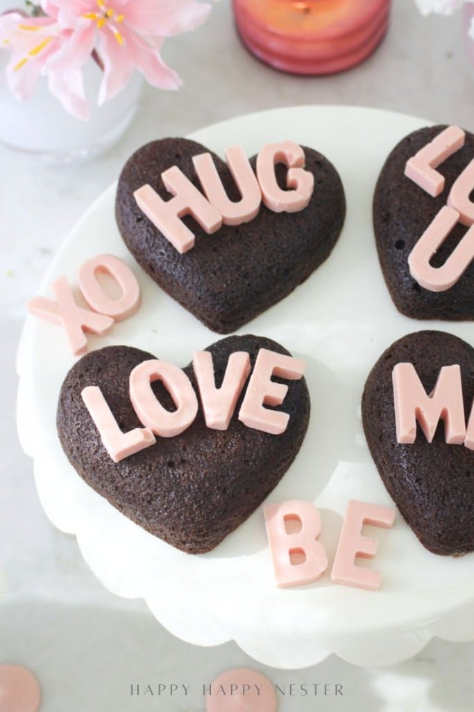 Heart-shaped chocolate cakes, inspired by a heart shaped brownies recipe, spell out phrases like "LOVE," "HUG," and "XO" in pink letters, all arranged on a white cake stand with a pink flower nearby.
