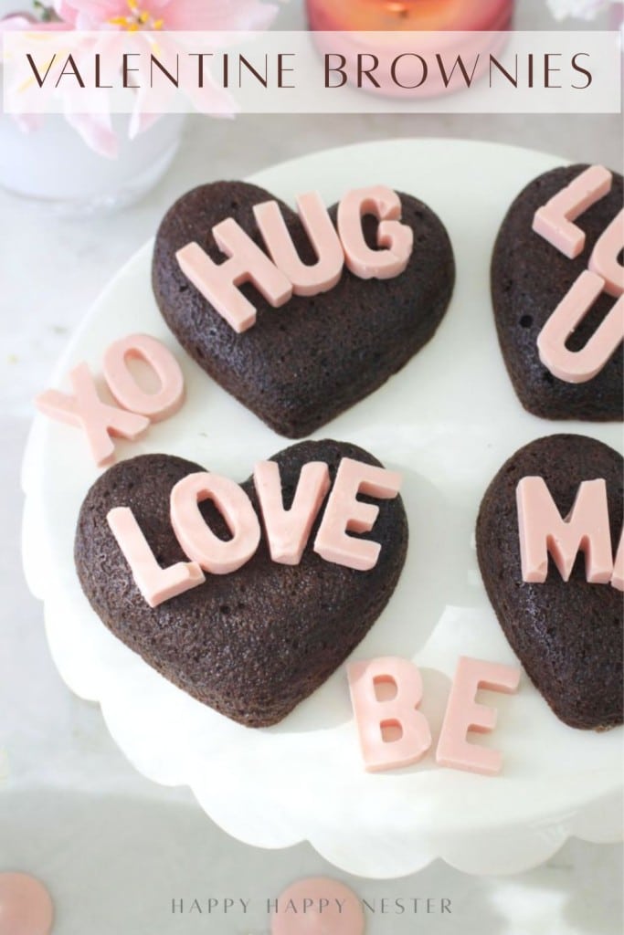 Heart-shaped brownies topped with pink letters spelling words like “HUG,” “LOVE,” “XO,” “BE,” and more, displayed on a white cake stand with “Valentine Brownies” text above.