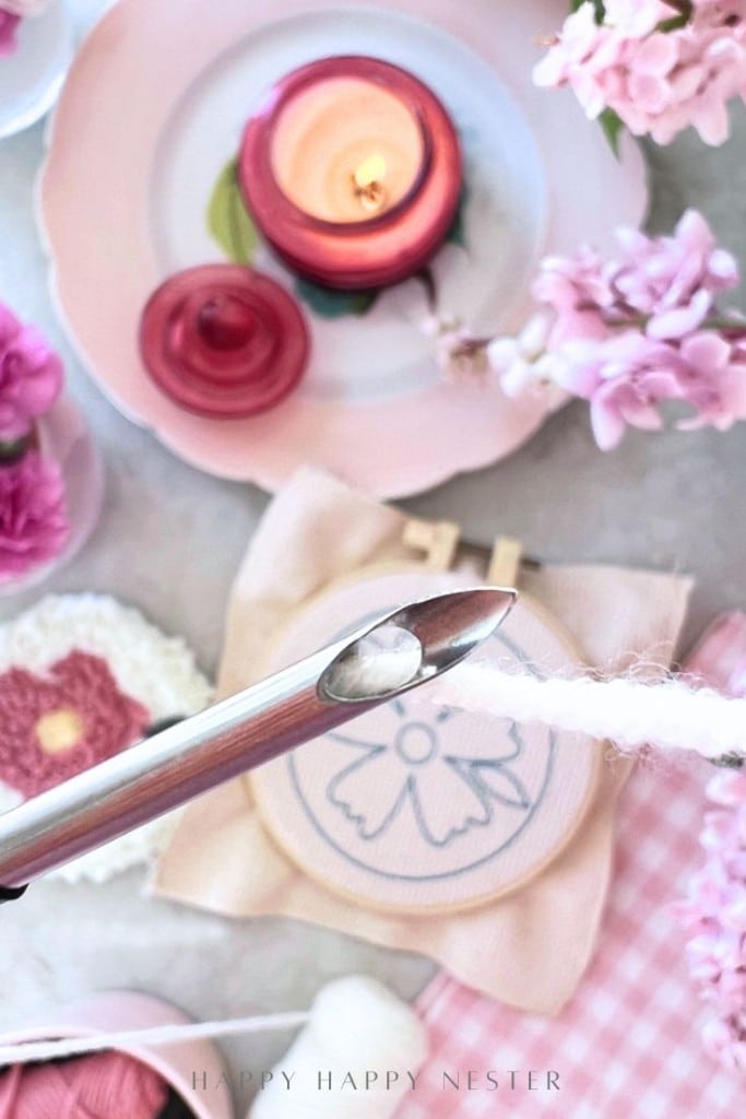 A close-up of a large needle threading white yarn, with pink flowers, a lit pink candle, embroidery hoop, crochet pieces, and pink textile items in the background, creating a cozy crafting scene.