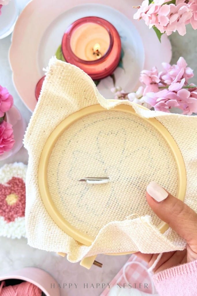 A hand holds an embroidery hoop with fabric, a needle, and a faint heart pattern. Pink flowers, yarn, and a lit candle are arranged in the background, creating a cozy crafting scene.