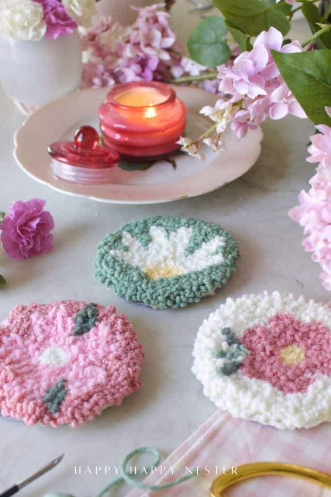 Three handmade floral coasters created with punch needle crafting sit on a table with pink flowers, a red candle in a glass holder, and a pink plate, creating a cozy and artistic spring-themed scene.