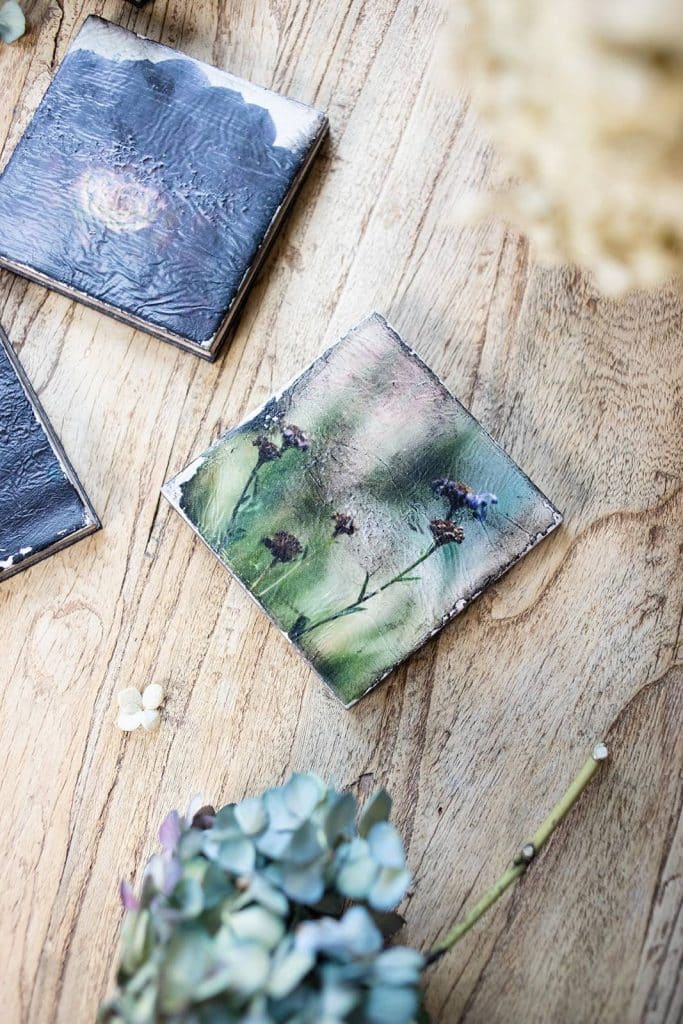Two handcrafted ceramic tiles with artistic floral and abstract designs rest on a light wooden surface, surrounded by dried flowers and small white petals—a charming addition to any crafting or DIY space.
