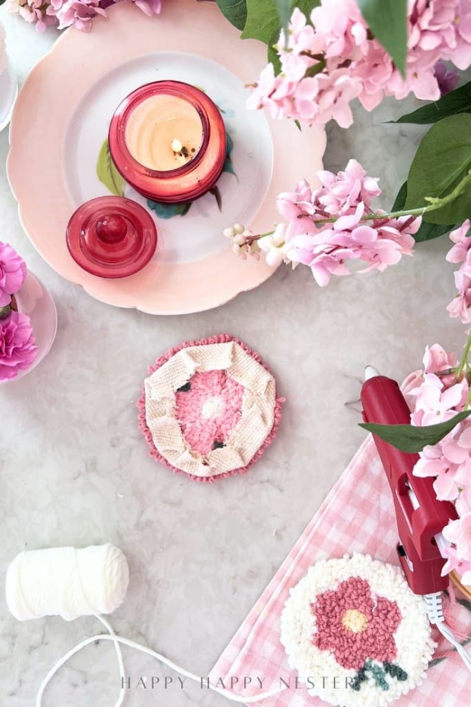 A table with pink flowers, a candle on a pink plate, a glue gun, white string, and two handmade round flower crafts on a pink gingham cloth. The scene is bright and arranged for a DIY project.