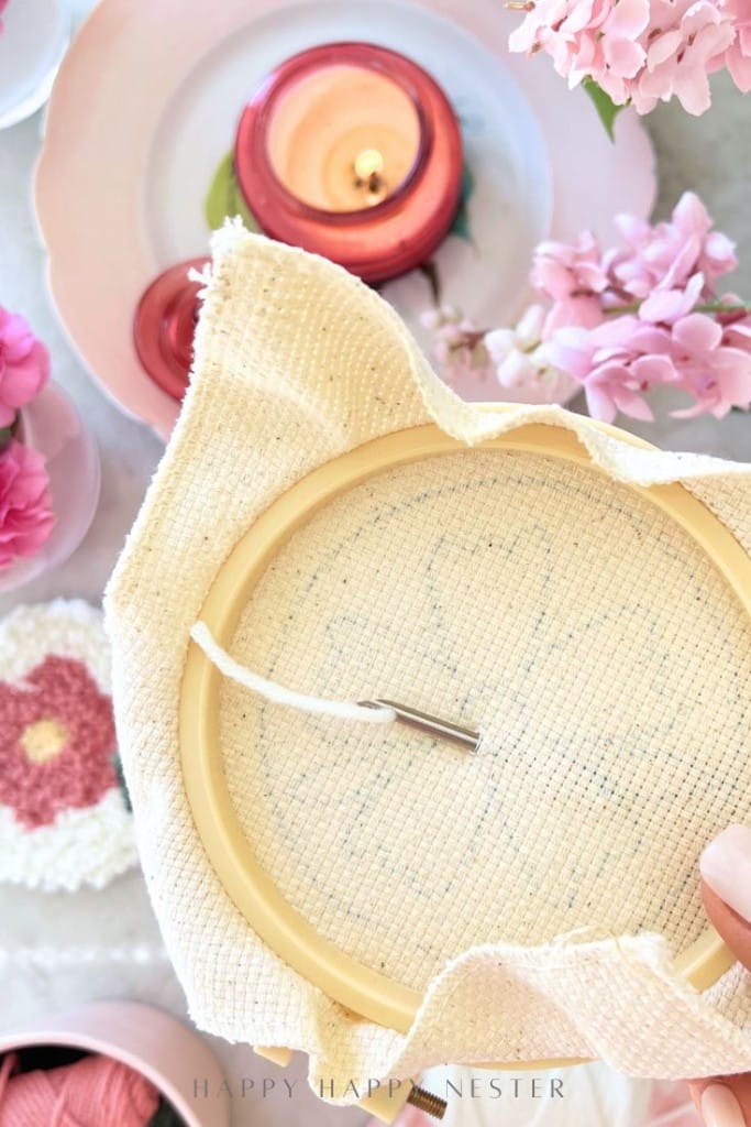 A hand holds an embroidery hoop with beige fabric, surrounded by pink flowers, a lit candle, and crochet items on a table. The needle and thread for punch needle crafting are visible on the fabric.