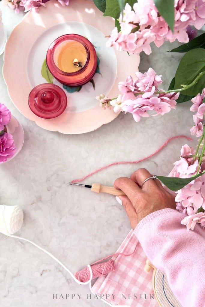 A hand with a crochet hook works with pink yarn on a marble surface, surrounded by pink flowers, a pink candle in a glass jar, and a pink gingham pouch. The bright, cozy scene invites you to explore DIY punch needle crafting.