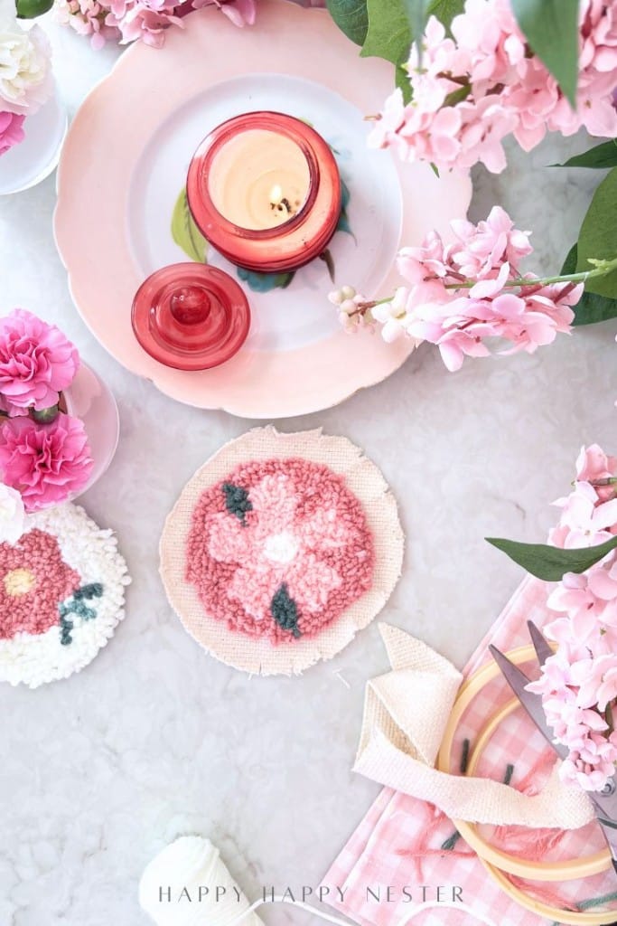 A top view of a pink-themed table with a candle, pink flowers, punch needle crafting DIY coasters, embroidery hoops, and a pink checkered cloth arranged on a white surface.