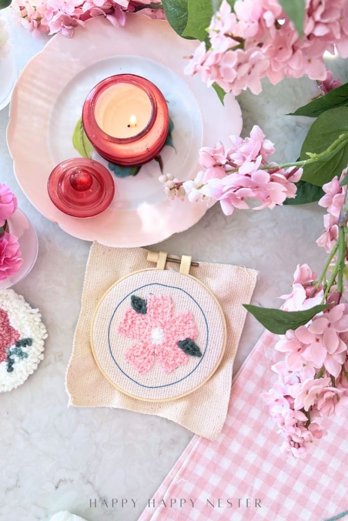A pink candle, cherry blossoms, a cup of tea, and a pink punch needle crafting embroidery in a hoop are arranged on a white surface with a pink gingham napkin, creating a soft, spring-themed DIY setting.