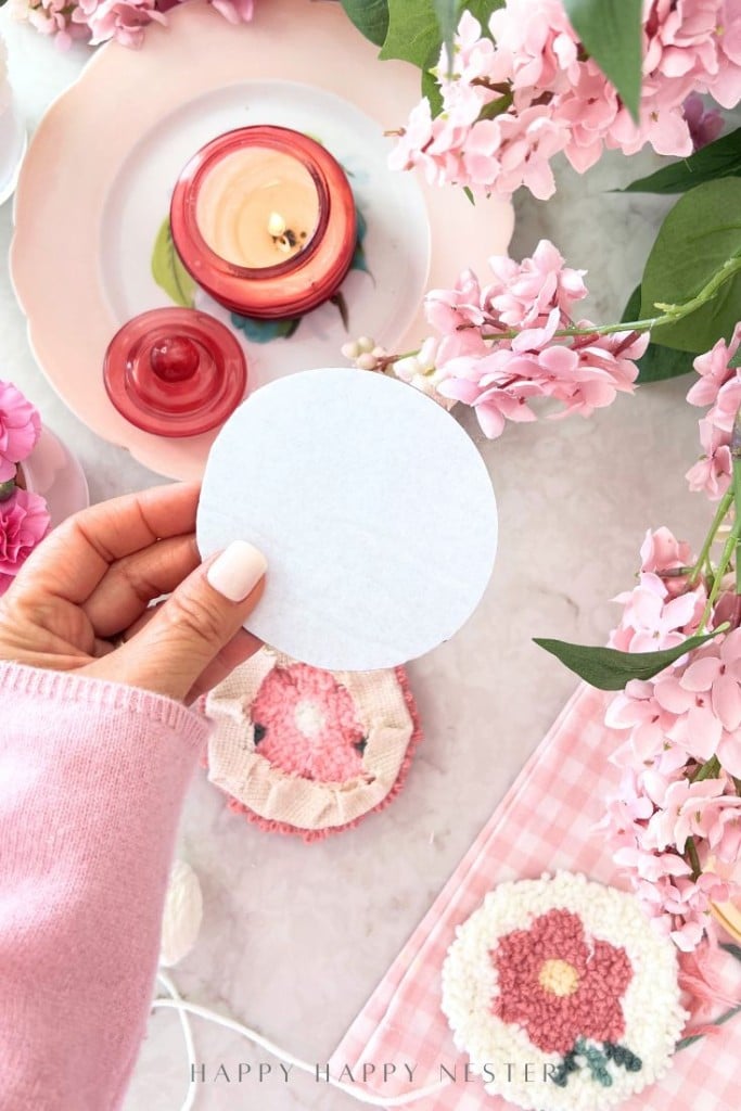 A hand holding a round white paper above a pink table setting with flowers, a candle, a pink gingham napkin, and floral coasters. The scene has a soft, springtime feel—perfect inspiration for punch needle crafting DIY projects.